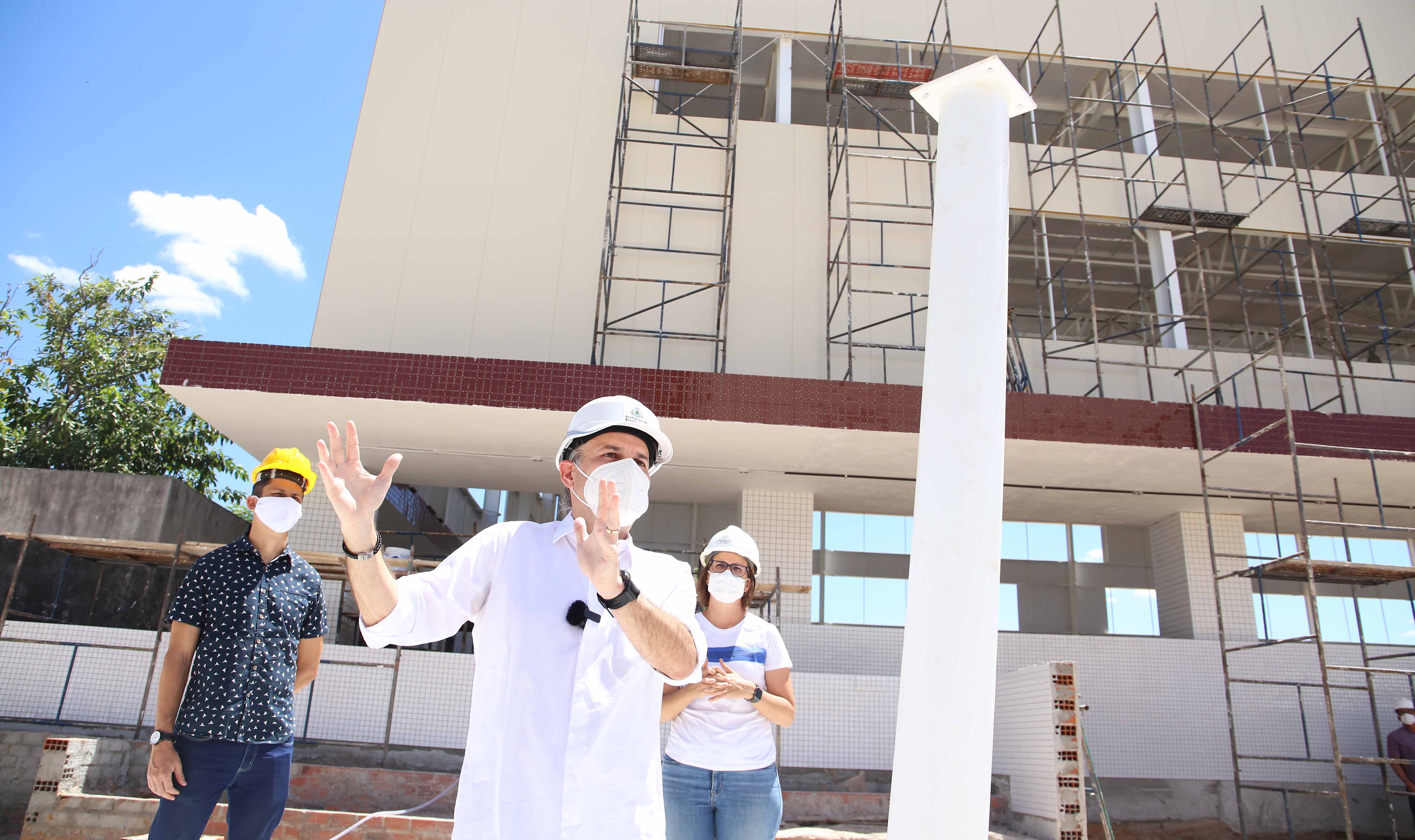 prefeito com coordenador da juventude e secretária Manuela Nogueira em pé posando para a foto, usando máscara e capacete de obras com prédio em construção ao fundo